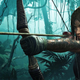 Lara Croft aims a large arrow using a wooden bow deep in a jungle.